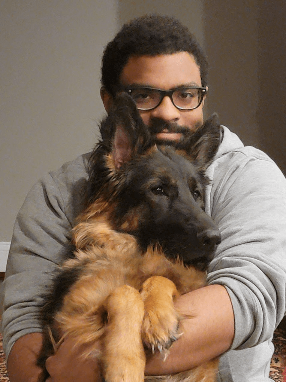 Elías Ramos, a young man with light brown skin, black-framed glasses, and a short mustache and goatee, dressed in a grey hooded sweatshirt and cradling a German shepherd puppy, Adi, in his arms.